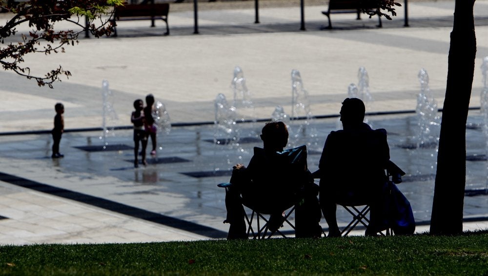 Dos personas a la sombra, mientras los niños se refresca en una fuente