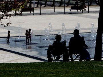 Dos personas a la sombra, mientras los niños se refresca en una fuente