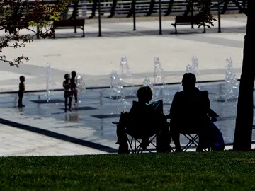 Dos personas a la sombra, mientras los niños se refresca en una fuente Dos personas a la sombra, mientras los niños se refresca en una fuente