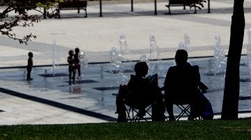 Dos personas a la sombra, mientras los niños se refresca en una fuente