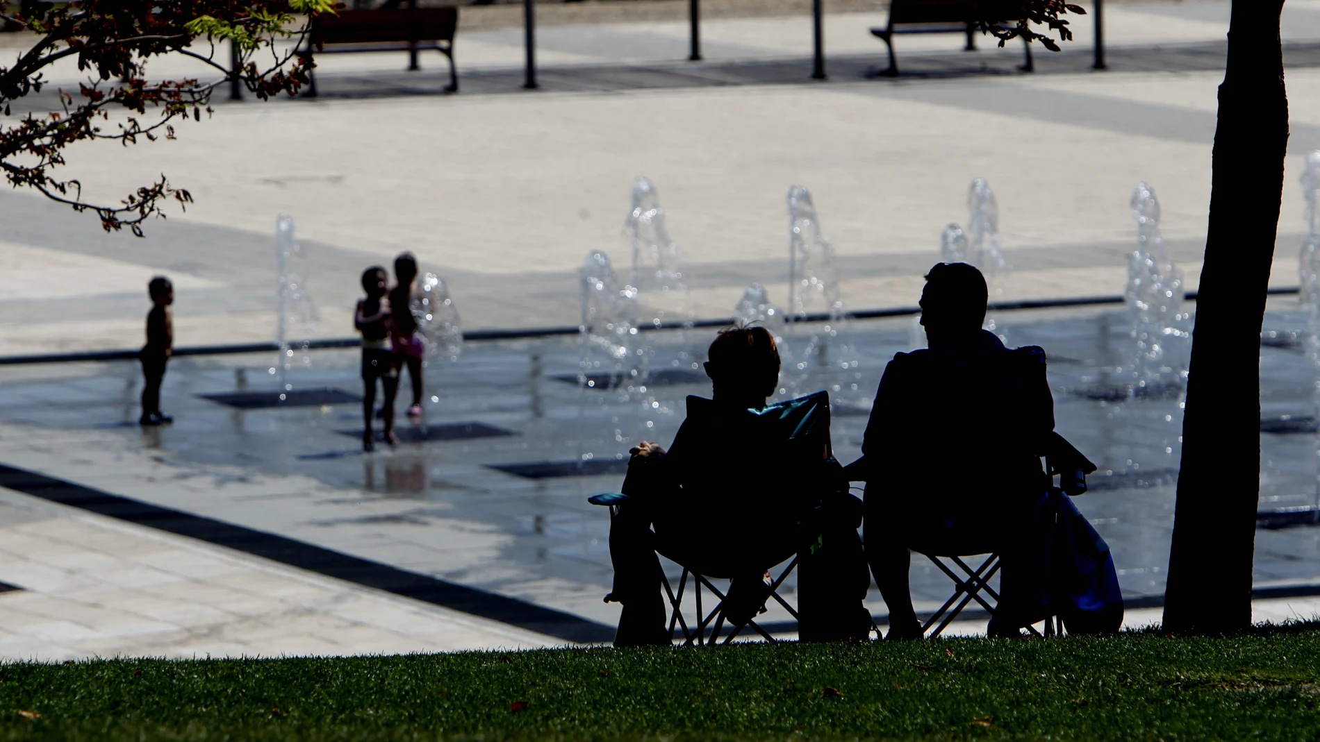 Dos personas a la sombra, mientras los niños se refresca en una fuente Dos personas a la sombra, mientras los niños se refresca en una fuente