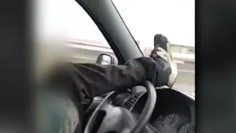 Dos jóvenes se graban conduciendo en dirección contraria y manejando el volante con el pie por una carrtera de Astillero Dos jóvenes se graban conduciendo en dirección contraria y manejando el volante con el pie por una carrtera de Astillero