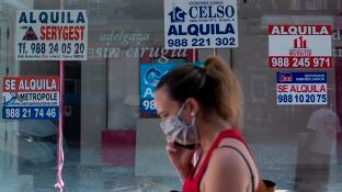 Una mujer con mascarilla pasa ante un comercio cerrado con varios carteles que anuncian su alquiler