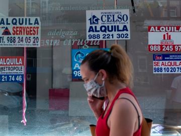 Una mujer con mascarilla pasa ante un comercio cerrado con varios carteles que anuncian su alquiler