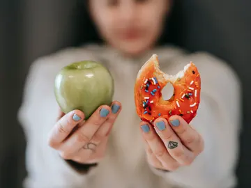 Una mujer sostiene un alimento saludable en una mano y, en la otra, un dulce poco saludable. Una mujer sostiene un alimento saludable en una mano y, en la otra, un dulce poco saludable.