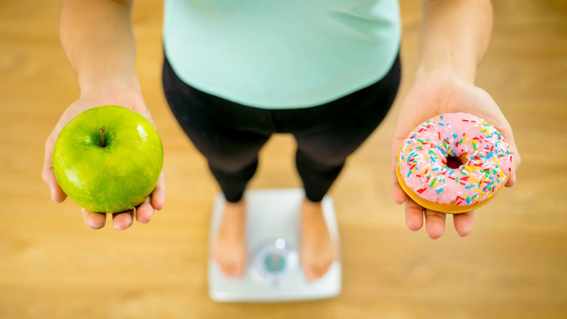 Chica subida a una báscula con una manzana en una mano y un dulce en la otra.