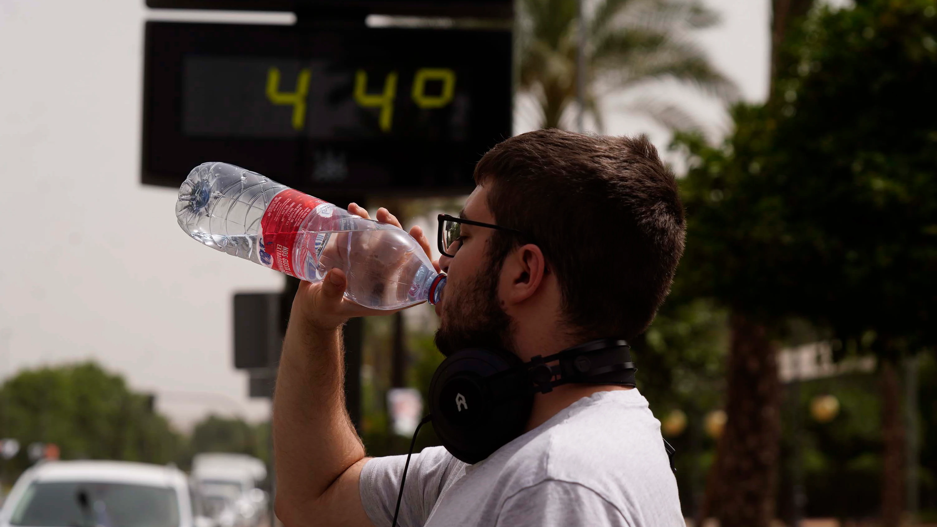 Un joven bebiendo agua frente a un termómetro con altas temperaturas Un joven bebiendo agua frente a un termómetro con altas temperaturas