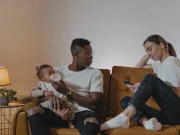 Bebé alimentándose con biberón en brazos de su padre, sentado en el sofá junto a su madre que consulta el teléfono. Bebé alimentándose con biberón en brazos de su padre, sentado en el sofá junto a su madre que consulta el teléfono.