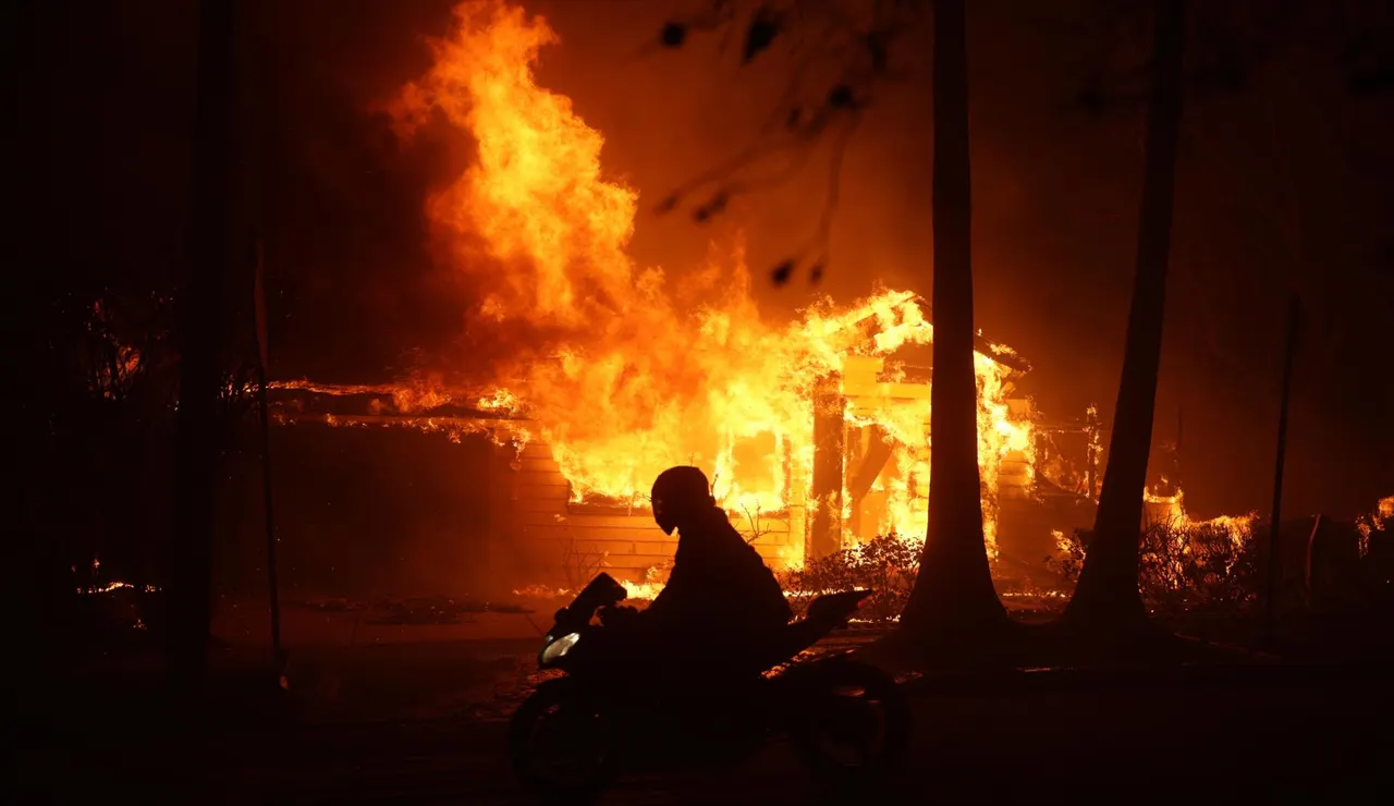 Una persona conduce su motocicleta junto a una casa en llamas durante los incendios en California
