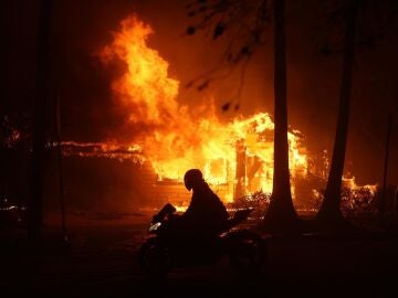 Una persona conduce su motocicleta junto a una casa en llamas durante los incendios en California