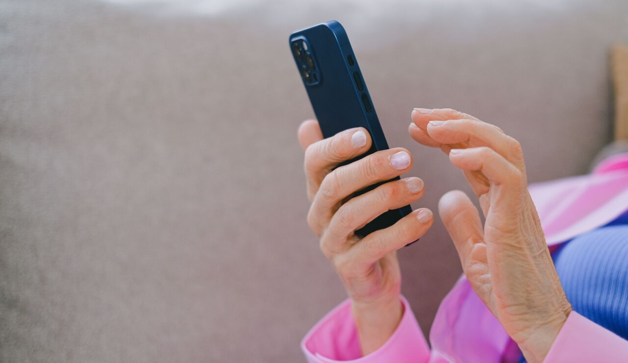 Un adulto mayor con chaqueta rosa usando un smartphone mientras se relaja en un sof&aacute; en interiores