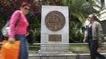 Varios viandantes frente a un monumento que hace honor a la antigua moneda griega