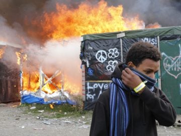 Un migrante se protege el rostro al pasar junto a chabolas en llamas durante el desmantelamiento del campamento conocido como la 'Jungla' de Calais 