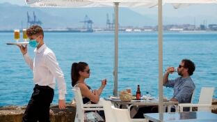 Un camarero con mascarilla pasa ante una pareja que almuerza en un restaurante de M&aacute;laga