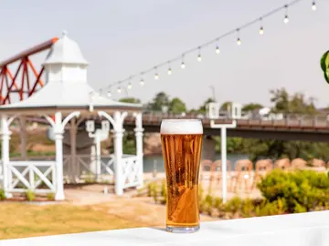 Copa de cerveza sobre una mesa. Al fondo vistas de una glorieta y un puente sobre un río Copa de cerveza sobre una mesa. Al fondo vistas de una glorieta y un puente sobre un río