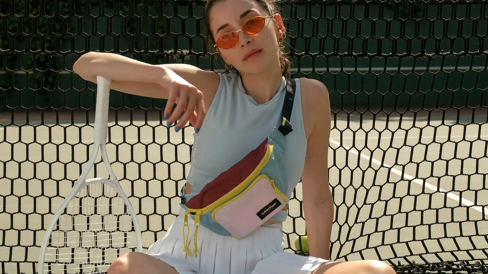 Chica con gafas de sol, riñonera, camiseta sin mangas, minifalda plisada, calcetines altos y deportivas en una pista de tenis.