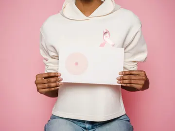 Mujer que sostiene entre las manos un dibujo de un pecho mientras luce un lazo contra el cáncer de mama. Mujer que sostiene entre las manos un dibujo de un pecho mientras luce un lazo contra el cáncer de mama.