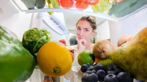Una mujer pensativa frente a la nevera, llena de verduras y frutas Una mujer pensativa frente a la nevera, llena de verduras y frutas